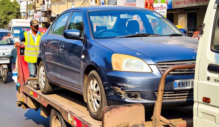 A damaged blue car being towed on a flatbed truck in a busy street of Aurangabad, showcasing professional towing services in Aurangabad with a worker assisting the process.